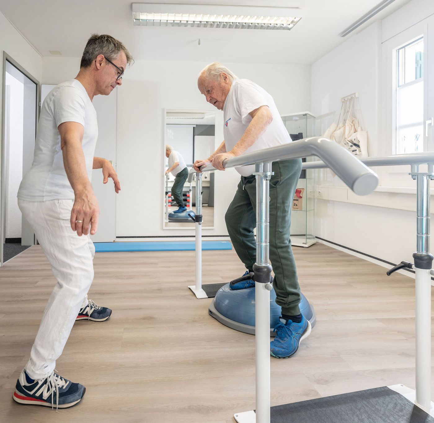 Physiotherapeut Manolito van Haren betreut älteren Patienten beim Gleichgewichtstraining auf dem Bosu-Ball in Physio Kinetic